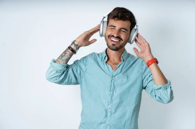 Beyaz arka planda turkuaz tişört giyen yakışıklı genç adam gülümsüyor. Kablosuz kulaklıkla en sevdiği müziği dinlediği için çok mutlu. Gözlerini kapatıyor..