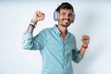 Beyaz arka planda turkuaz gömlek giyen kaygısız yakışıklı genç adam, dişlek gülüşü ile kol dansı yapıyor, tasasız hareketler, müziğin ritmiyle müzik listesinden kulaklıkla müzik dinliyor.