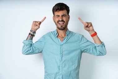 Beyaz arka planda turkuaz gömlekli çılgın yakışıklı bir genç adam fotoğrafı. Çığlık atıyor ve parmakları saçları kapalı gözleri işaret ediyor.