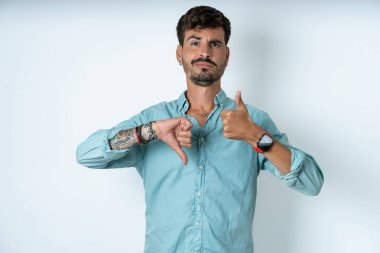 Beyaz arka planda turkuaz gömlek giyen yakışıklı genç adam baş parmağını aşağı işareti gösteriyor.