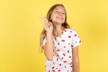 Sarı arka planda puantiyeli tişört giyen beyaz kız kameraya bakıp gülümsüyor ve zafer işareti yapıyor. İki numara..