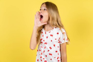 caucasian kid girl wearing polka dot shirt over yellow background shouting and screaming loud to side with hand on mouth. Communication concept. clipart