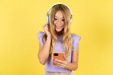 Sarı arka plan üzerine mor tişört giyen mutlu sarışın kız akıllı telefon ekranında odaklanırken iyi hissediyor. İnsanlar, teknoloji, yaşam tarzı