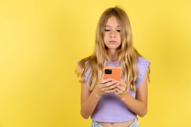 Sarı arkaplan üzerine mor tişört giyen odaklanmış sarışın kız sosyal medya haberlerini ya da önemli e-postaları okuyarak akıllı telefon kullan