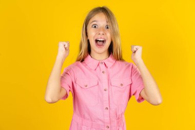 Şok olmuş pembe ceketli kız sarı arka planda şans piyangosunu kazan elini kaldır ve bağır