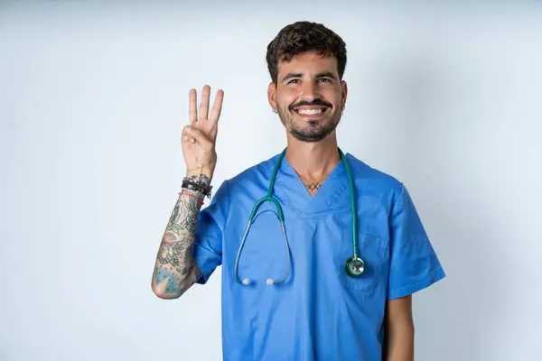 英俊的护士 身穿外科制服 背景洁白 手指头指向三号 面带微笑 自信而快乐 — 图库照片