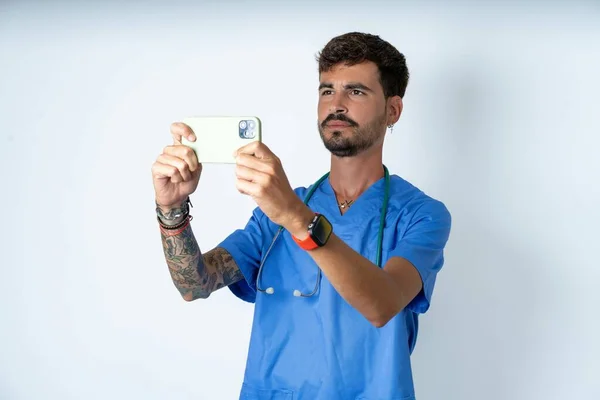 handsome nurse man wearing surgeon uniform over white background taking a selfie to post it on social media or having a video call with friends.