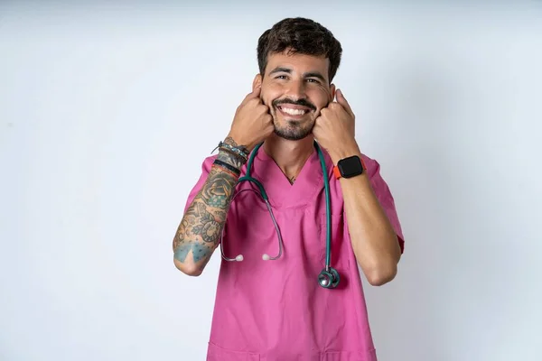 Happy handsome nurse man wearing surgeon uniform over white background keeps fists on cheeks smiles broadly and has positive expression being in good mood