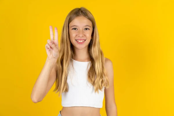 黄色の背景に白いTシャツを着て 自信と幸せを笑いながら指ナンバー2を指で指で指を指さすブロンドの少女 — ストック写真