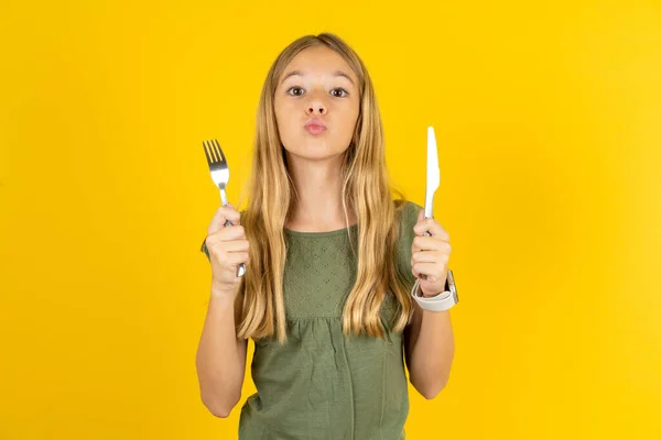 hungry blond little girl wearing khaki blouse over yellow background holding in hand fork knife want tasty yummy pizza pie