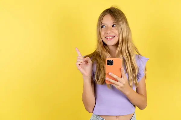 Улыбающаяся Блондинка Фиолетовой Футболке Жёлтом Фоне Держит Мобильный Телефон Указывает — стоковое фото