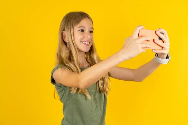 blond little girl wearing khaki blouse over yellow background taking a selfie to post it on social media or having a video call with friends.