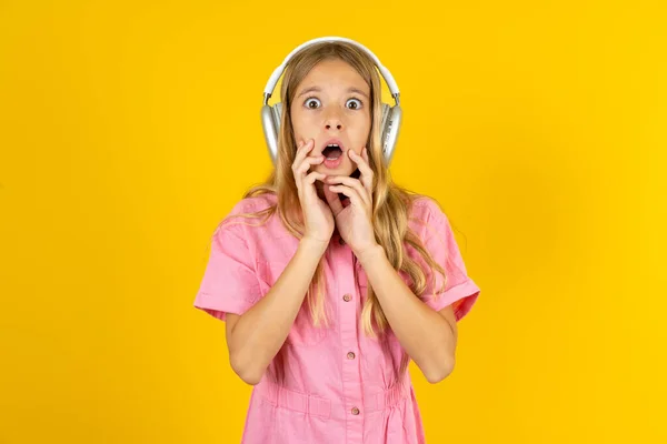 穿着粉色夹克 披着黄色背景的惊慌失措的女孩在镜头前不敢张大嘴 耳朵上戴着无线立体声耳机 — 图库照片