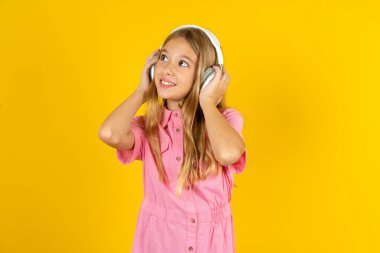 Sarı arka planda pembe ceket giyen kız stereo kulaklık takıyor müzik dinliyor. İnsanlar yaşam tarzı kavramını hobiliyor.