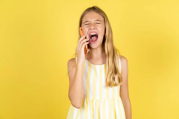 Overemotive happy girl wearing yellow dress over yellow background laughs out positively hears funny story from friend during telephone conversation