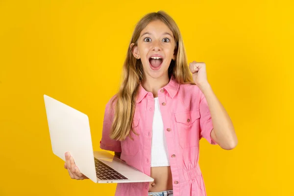 在黄色背景下穿着粉色夹克的女孩拿着电脑张开嘴举起拳头 — 图库照片