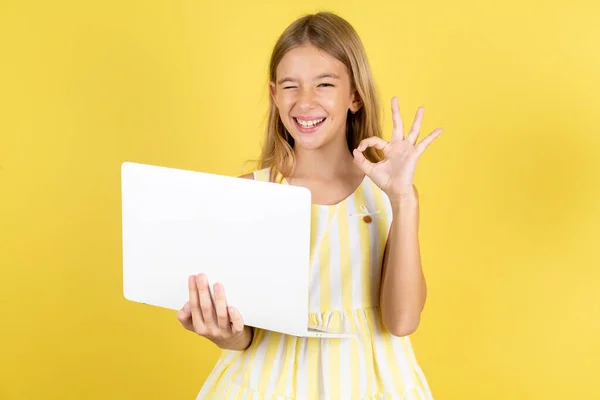 用显示Ok Sign Winkin的笔记本电脑在黄色背景下穿着黄色衣服的迷人的 快乐的 熟练的女孩 — 图库照片