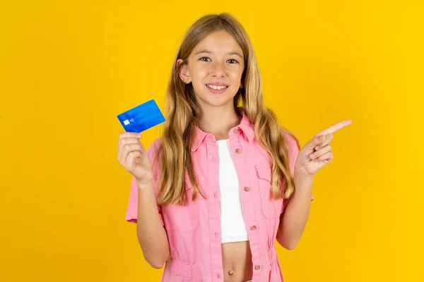 stock image Smiling girl wearing pink jacket over yellow background showing debit card pointing finger empty space