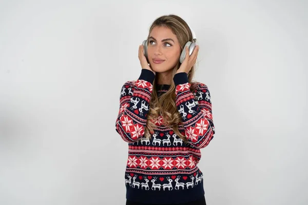 漂亮的金发女人穿着针织圣诞毛衣 戴着立体声耳机 聚精会神地听音乐 人们的业余爱好生活方式概念 — 图库照片