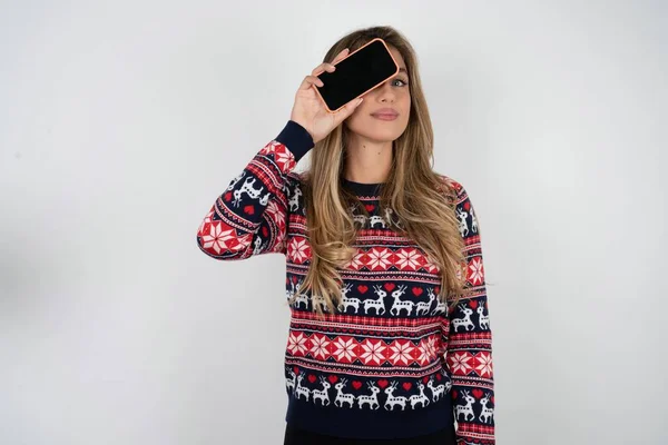 漂亮的金发女人 头戴针织圣诞毛衣 手持现代智能手机 一边微笑 一边捂住一只眼睛 — 图库照片