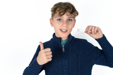 Mavi kazaklı yakışıklı beyaz çocuk görünmez bir diş teli tutuyor ve baş parmağını kaldırıyor. Bu yeni tedaviyi öneriyor. Diş sağlığı konsepti.