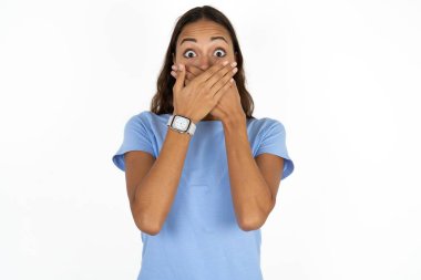 Beyaz arka plan üzerine mavi tişört giyen genç ve güzel İspanyol kadın yanlışlıkla elleri ile ağzını kapatırken şok oldu. Gizli konsept.