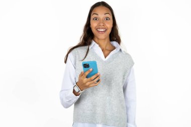 Beyaz arka plandaki genç, güzel İspanyol kadın cep telefonunu elinde tutuyor ve olumlu haberler veriyor, modern cep telefonu kullanıyor.