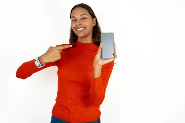 白い背景の上に赤いタートルネックを着ている若いヒスパニック女性が笑顔でコピースペースをモックします 空白のスクリーンが付いている携帯電話の指の指を指すこと — ストック写真