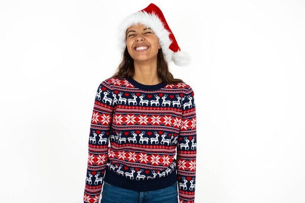 Positive beautiful hispanic woman wearing knitted sweater and santa claus hat over white background with overjoyed expression closes eyes and laughs shows white perfect teeth