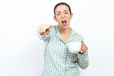 Yeşil ekose pijama giyen, elinde bir kupa tutan, kameraya kızgın ve sinirli, sizinle kavga etmeye hazır genç bir kadın..