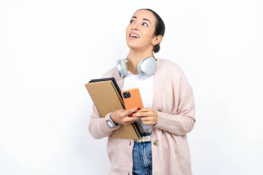 Güzel bir Arap kız öğrenci beyaz arka plan üzerinde defterler taşıyor cep telefonunu tutuyor ve rüya gibi bir bakışla yana bakıyor..