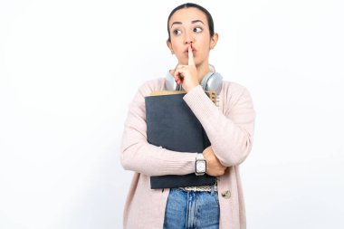 Güzel Arap kız öğrenci beyaz arka planda not defterleri taşıyor. Sessiz jest, işaret parmağını dudaklara doğru tutuyor. Sessiz ol işareti yapıyor. Sırrımızı paylaşmamamızı istiyor.