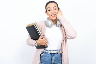 Mutlu, güzel bir Arap kız öğrenci beyaz arka planda defterler taşıyor. Yumruklarını yanaklarında gezdiriyor. Gülümsüyor ve olumlu bir ifade sergiliyor.