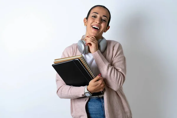 beautiful arab woman student carries notebooks over white background laughs happily keeps hand on chin expresses positive emotions smiles broadly has carefree expression