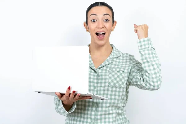 身穿绿色格子布睡衣 头戴白色背景的年轻貌美的女子举着计算机张开的嘴举起拳头 — 图库照片