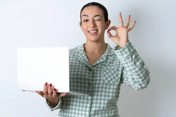 在白色背景上穿着绿色格子花睡衣的年轻貌美的女士手持无线上网本手指头显示出Okey符号 — 图库照片