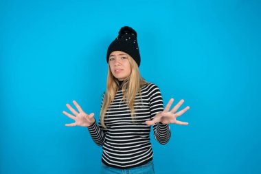 Güzel bir kız çocuğu örülmüş siyah şapka ve mavi arka planda çizgili balıkçı yaka giyiyor korku ifadesinden korkuyor ve dehşete düşüyor. El hareketlerini durdur, şok içinde bağır. Panik konsepti.