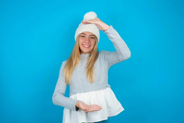 白い編まれた帽子と青いセーターのジェスチャーを身に着けている美しい少女は 大きくて大きいサインを示し シンボルを測定します カメラを見ている笑顔 — ストック写真