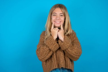 Kahverengi örgü süveter giyen, dişlek gülüşlü, işaret parmaklarını ağzının yanında tutan, parmaklarını işaret eden, neşeli gülümsemeye zorlayan mutlu kız.