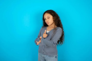 Neşeli güzel kız mavi arka planda sıradan bir ceket giyiyor ve işaret parmaklarıyla kameraya işaret ediyor, birini seçiyor, parmak tabancası yapıyor..