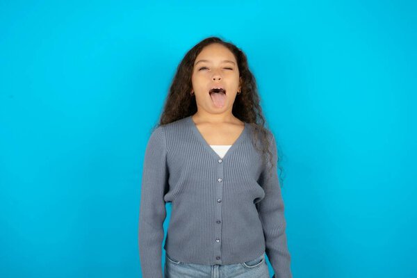 Beautiful kid girl wearing casual jacket over blue background sticking tongue out happy with funny expression. Emotion concept.