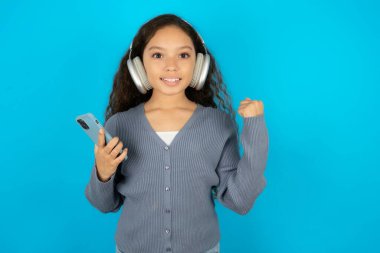 Mavi arka plan üzerine gündelik ceket giyen güzel bir kız çocuğu modern cep telefonunu kulaklıklara bağlar, iyi duygulardan yumruğunu sıkar, sevinçten haykırır.,