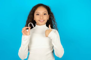 Mavi arka plan üzerine beyaz balıkçı yaka giyen güzel bir kız. Görünmez bir diş teli tutuyor ve baş parmağını kaldırıyor. Bu yeni tedaviyi öneriyor. Diş sağlığı konsepti.