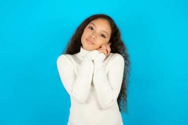 Büyüleyici, ciddi, beyaz balıkçı yaka kazak giyen güzel bir kız çocuğu. Mavi arka planda elleri kameraya nazikçe gülümsüyor.