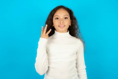 Mavi arka planda beyaz balıkçı yakalı güzel bir kız gülümsüyor ve arkadaş canlısı görünüyor, üç ya da üç numarayı gösteriyor, geriye doğru sayıyor.