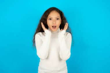 Şaşırmış mutlu, mavi arka planda beyaz boğazlı kazak giyen güzel bir kız. Giysilerde büyük indirimler görmekten memnun. Şok olduğunu ifade ediyor..