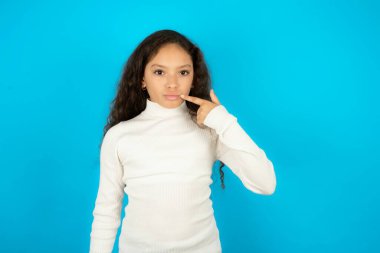 Beyaz balıkçı yakalı, mavi arka planda güzel bir kız çocuğu işaret ederek dudaklarını işaret ediyor, ameliyattan sonra etkisini gösteriyor.,