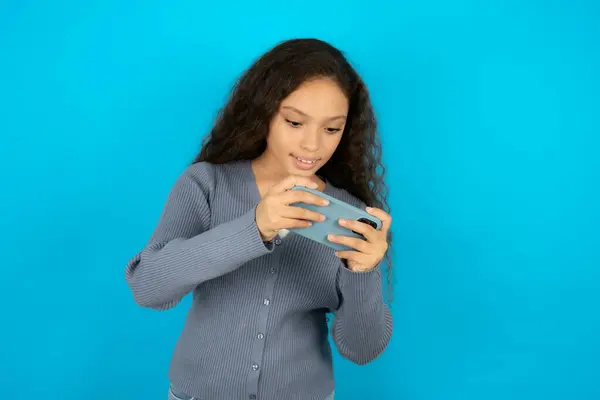 ビデオゲームやチャットをする手のセルで保持ブルーの背景の上にカジュアルなジャケットを着ている美しい子供の少女 — ストック写真