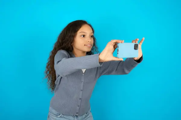 ソーシャルメディアに投稿するか 友人とビデオ通話をするためにセルフィーを取る青い背景の上にカジュアルなジャケットを着ている美しい子供の少女 — ストック写真