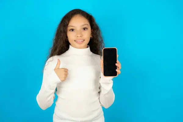 青い背景に白いタートルネックを身に着けている美しい子供の少女 空白のスクリーン スマートフォンを表示 親指で新しいアプリをお勧めします — ストック写真